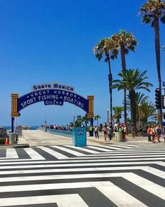 people are walking on the beach near some palm trees and a blue sign that says santa monica