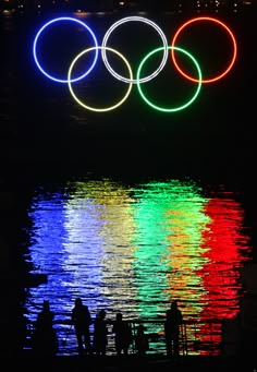 the olympic rings are lit up at night over water with people standing in front of it