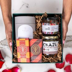 a woman's hands holding an open gift box filled with food and candles, surrounded by rose petals