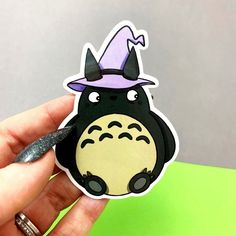 a hand holding a sticker with a totoro wearing a purple hat on it