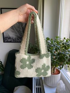 a hand holding a green and white purse next to a potted plant