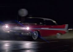 an old red car driving down the road at night with fog coming from behind it