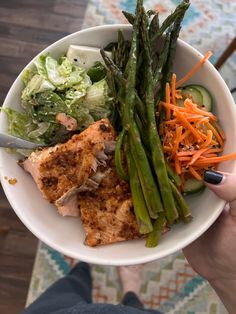 a person holding a bowl of food with asparagus, carrots and meat