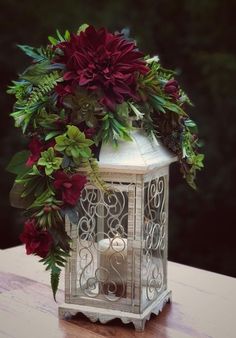 a white lantern with red flowers and greenery in it sitting on a wooden table