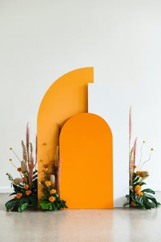 an orange and white arch with flowers on the floor in front of it that is made out of cardboard