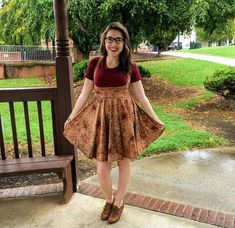 a woman standing in front of a wooden bench wearing glasses and a dress with floral print