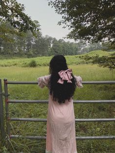 a woman standing in front of a fence looking out at the grass and trees behind her