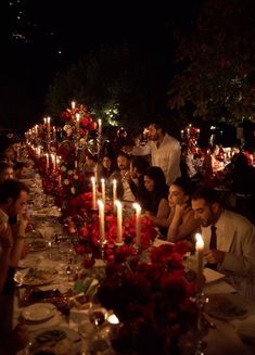 a group of people sitting at a long table with candles in front of them and flowers on the table