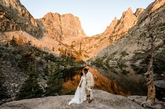 a bride and groom standing on the edge of a cliff overlooking a mountain lake at sunset