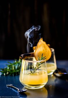 two glasses filled with liquid sitting on top of a table next to spoons and herbs