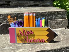 pencils and markers in a wooden box with the word miss written on it next to some rocks