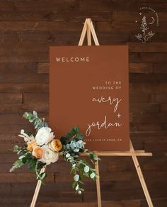 a welcome sign with flowers and greenery sits on an easel in front of a wooden wall