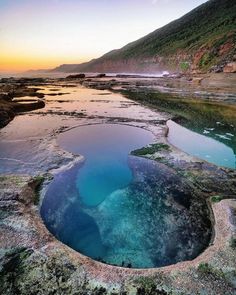 the blue pool is surrounded by large rocks and green vegetation, as the sun sets in the distance