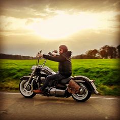 a man riding on the back of a motorcycle down a road next to a lush green field