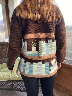 a woman standing in front of a window with her back turned to the camera, wearing a brown and blue crocheted sweater