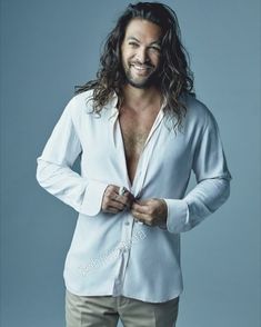 a man with long hair standing in front of a blue background wearing pants and a white shirt