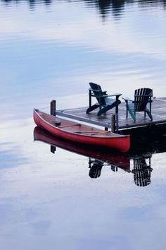 a red boat sitting on top of a lake next to a dock with two chairs