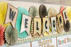 a welcome sign with paper fans hanging from it's sides in front of a bulletin board