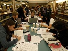 several people are sitting at a table with papers and laptops in a library while others look on