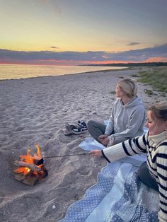 two women sitting on the beach next to a campfire