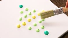 someone using a paintbrush to decorate small green and yellow drops on a white paper