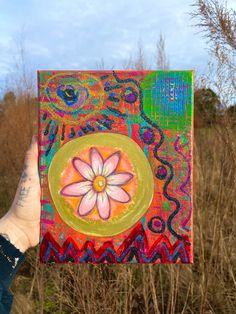 a person holding up a colorful painting in front of some grass and trees with the sky behind them