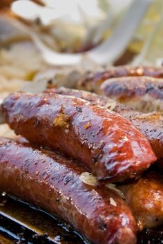 two sausages on a grill with onions and other food items in the back ground