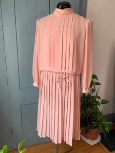 "This lovely vintage dress would be perfect for a spring party! It is peach crepe with satin yoke, collar, cuffs and belt (all polyester). It has a pleated front and skirt. It closes with to buttons at the neck and has buttons on the cuffs. It is long sleeved and the skirt is about mid-calf length. It is marked Mavinette size 14. The bust is approximately 42\" and the waist is approximately 38\" it is in good condition with just a little spot on the front of the skirt.  (it may come out with some spot treatment I haven't tried). Please see pictures." Pink Pleated Skirt Dress For Formal Occasions, Pink Pleated Formal Dress, Pink Formal Dress With Pleated Waist, Formal Pink Pleated Dress, Spring Party Vintage Pleated Dress, Classic Vintage Dress For Spring Party, Vintage Pleated Dress For Party, Pleated Vintage Dress For Party, Classic Vintage Dress For Spring Formal