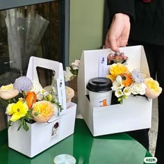 two white boxes with flowers in them sitting on a green counter top next to a person holding a cup