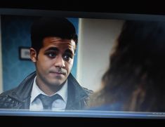 a man in a jacket and tie is looking at another person on the television screen