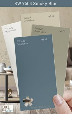 a hand holding three different shades of paint in front of a living room with the words sw 7604 smoky blue on it