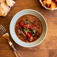 a bowl filled with stew next to a fork and knife on a wooden counter top