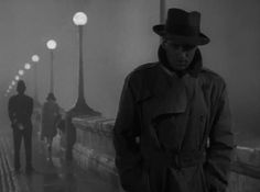 a man in a trench coat and hat standing on a bridge with people walking by