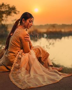 a woman sitting on the ground next to a body of water with her arms around her neck