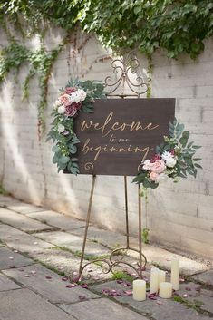 a welcome sign with flowers and greenery on it next to candles in front of a brick wall