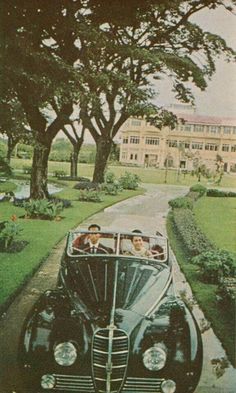 an old black car driving down a street next to a lush green park filled with trees