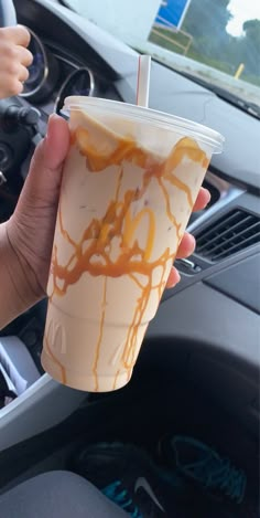 a hand holding a cup with caramel sauce on it in front of a steering wheel