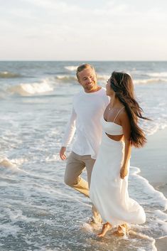 a man and woman are walking in the water at the beach, one is wearing a white dress