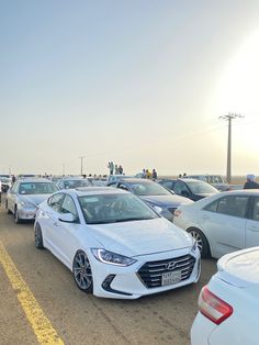 many cars are parked in a parking lot with people walking on the sidewalk and looking at them