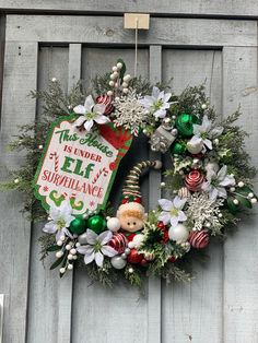 a christmas wreath hanging on the side of a wooden door with an elf's sign