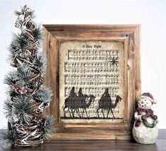 a snowman is sitting next to a christmas tree and a framed cross stitch pattern