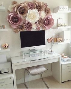 a white desk topped with a computer monitor next to a wall covered in paper flowers