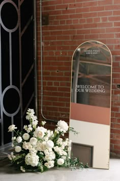 white flowers sitting on the ground in front of a welcome sign and brick wall behind it