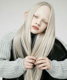 a woman with long blonde hair wearing a black jacket and grey sweater is holding her hands on her chest