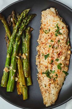 a black plate topped with fish and asparagus on top of a white table