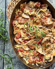 a skillet filled with mushrooms and tomatoes on top of a wooden table next to herbs