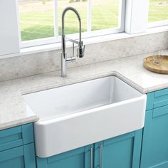 a white kitchen sink sitting under a window next to blue cabinets and counter top with a cutting board on it