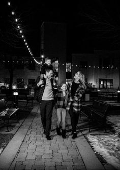 a man and woman walking down a sidewalk at night with lights strung up above them