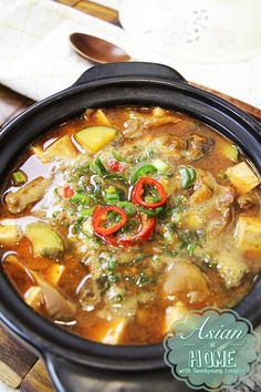 a black bowl filled with soup and vegetables on top of a wooden table next to a spoon