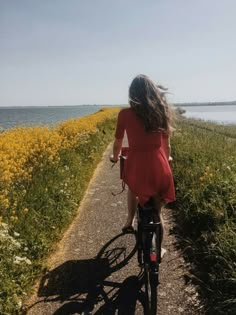 a woman in a red dress riding a bike on a path by the water with wildflowers